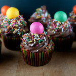 Chocolate malt cupcakes on a cutting board. Each cupcake is topped with gigantic malted milk balls, chocolate frosting, and vibrant sprinkles.