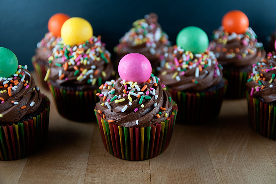 Chocolate malt cupcakes on a cutting board. Each cupcake is topped with gigantic malted milk balls, chocolate frosting, and vibrant sprinkles.